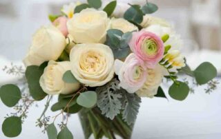 Wedding Table flowers of Ivory roses and delicate white peonies create a soft, ethereal base, while hints of blush pink ranunculus and fragrant freesia add a touch of warmth. The arrangements are complemented by trailing eucalyptus and silvery dusty miller, adding a graceful, flowing elegance