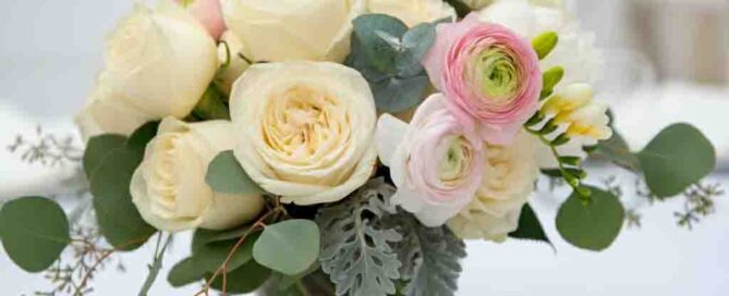 Wedding Table flowers of Ivory roses and delicate white peonies create a soft, ethereal base, while hints of blush pink ranunculus and fragrant freesia add a touch of warmth. The arrangements are complemented by trailing eucalyptus and silvery dusty miller, adding a graceful, flowing elegance